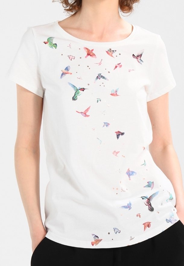 Chemise blanche à manches courtes avec de petits oiseaux colorés dispersés en diagonale sur le devant, portée par une personne les mains dans les poches.