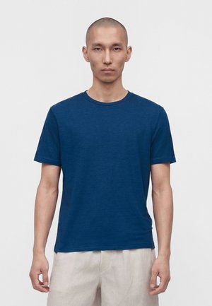 Hombre con cabeza rapada que lleva una camiseta azul lisa de manga corta y pantalones beige claro, de pie frente a un fondo blanco.