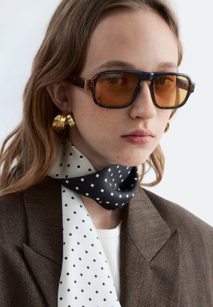 Jauna moteris dėvi didelio dydžio gintaro spalvos akinius, didelius auksinius žiedinius auskarus, rudą švarką ir juodai baltos spalvos raštuotą šaliką ant kaklo.