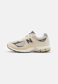 Baskets New Balance beige avec une tige en mesh et en daim, des accents gris, une semelle rembourrée et un logo N proéminent sur le côté.