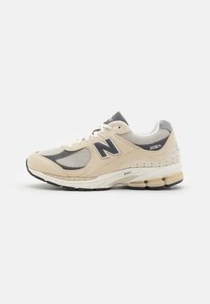 Baskets New Balance beige avec une tige en mesh et en daim, des accents gris, une semelle rembourrée et un logo N proéminent sur le côté.