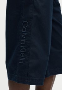 Κοντό παντελόνι σε ναυτικό μπλε χρώμα με το λογότυπο Calvin Klein κεντημένο κάθετα κατά μήκος της πλαϊνής ραφής, φορεμένο από ένα άτομο.