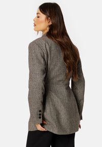 Blazer a motivo chevron in grigio e marrone, con spalle strutturate, taglio sartoriale e bottoni neri decorativi sulle maniche.