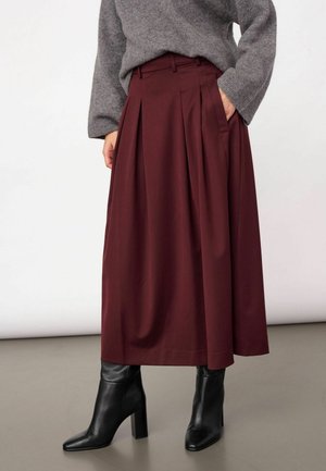 Femme portant une jupe midi plissée bordeaux, un pull gris et des bottines noires à talons, debout avec une main dans la poche.