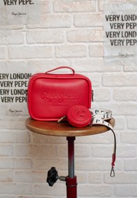 Borsa in pelle rossa con finitura testurizzata, logo impresso, portamonete rotondo e tracolla a fantasia su un tavolo di legno contro un muro di mattoni.