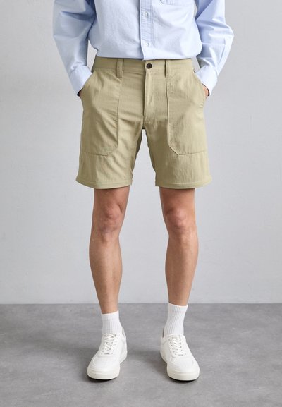 Shorts vert clair avec deux poches avant, en tissu de coton ; portés avec une chemise bleue et des baskets blanches, accompagnés de chaussettes blanches.