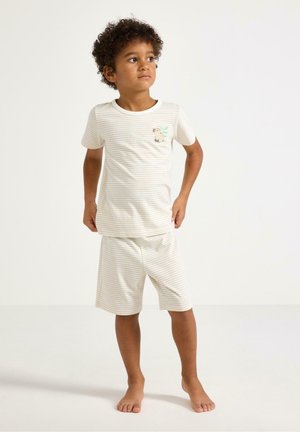 Chłopiec ubrany w beżowo-białą koszulkę w paski z naszywką psa oraz pasujące szorty, stojący boso na białym tle.