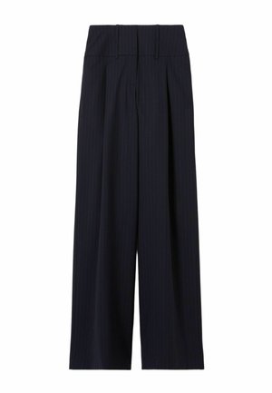 Femme portant un costume bleu marine à fines rayures avec un pantalon large, une chemise à rayures bleues, des chaussures marron et un pull marron à rayures posé sur les épaules, debout sur fond blanc.