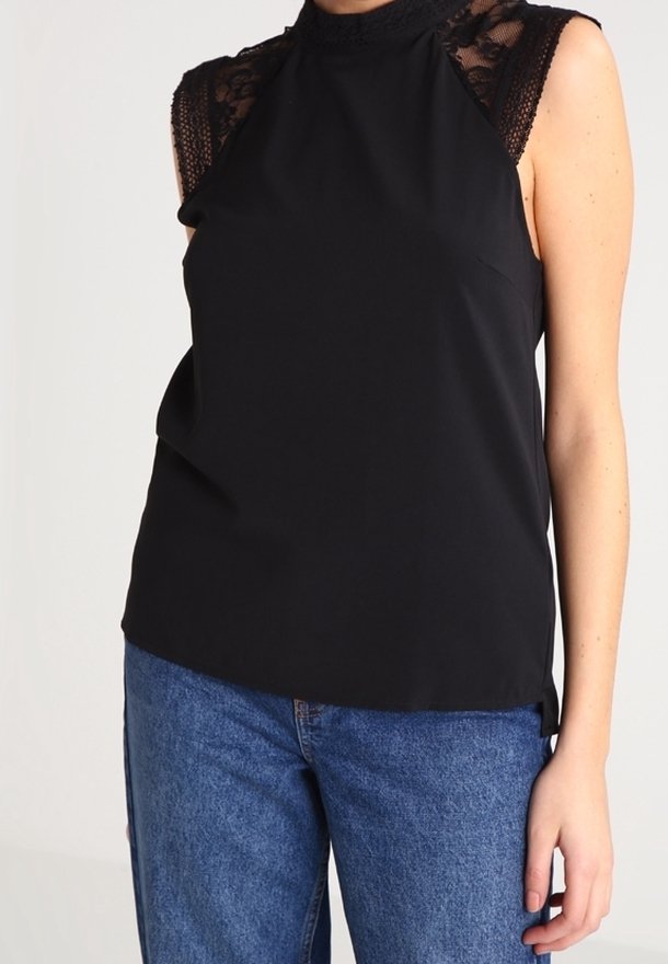 Femme portant un haut noir sans manches avec des épaules en dentelle, associé à un jean denim bleu sur un fond uni.