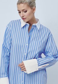 Ανοιχτό μπλε και λευκή ριγέ πουκάμισο με λευκό γιακά, μακριά μανίκια και τσέπη. Διαθέτει κουμπί κλεισίματος και αντίθετους μανσέτες.