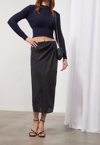 Pull tricoté court bleu marine avec des motifs complexes, associé à une longue jupe en satin noir. La mannequin porte des talons noirs à brides.