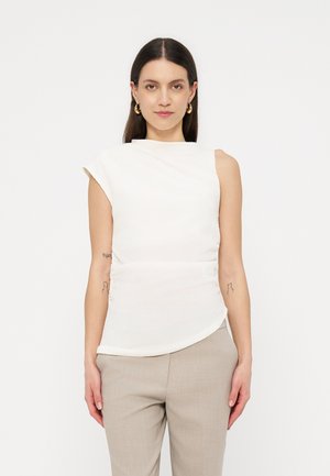 Γυναίκα που στέκεται μπροστά σε λευκό φόντο, φοράει λευκό αμάνικο τοπ, μπεζ παντελόνια, χρυσούς κρίκους στα αυτιά, με ορατά τατουάζ στα χέρια.