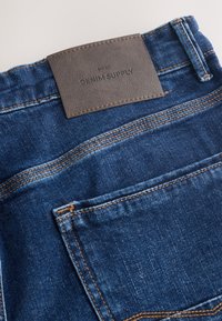 Jean en denim bleu montrant la poche arrière et un patch en cuir marron portant l’inscription « Next Denim Supply », cousu à la taille.