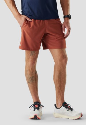 Homme portant un short de sport couleur rouille, des chaussures de course blanches avec des accents noirs et orange, et une montre noire au poignet, debout les mains détendues.