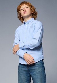 Camisa azul clara de botão com mangas longas, apresentando um pequeno logotipo no peito, emparelhada com jeans de denim azul. Textura do tecido suave.
