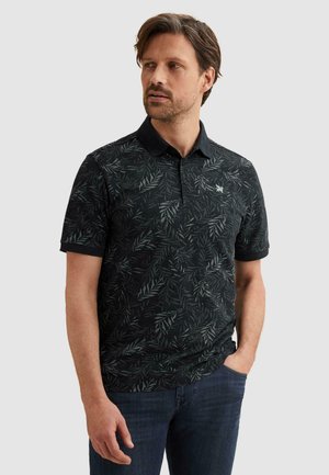 Mann trägt ein schwarzes Poloshirt mit kurzen Ärmeln und dunklem Blattmuster sowie blaue Jeans, steht mit einer Hand in der Tasche und schaut nach links.