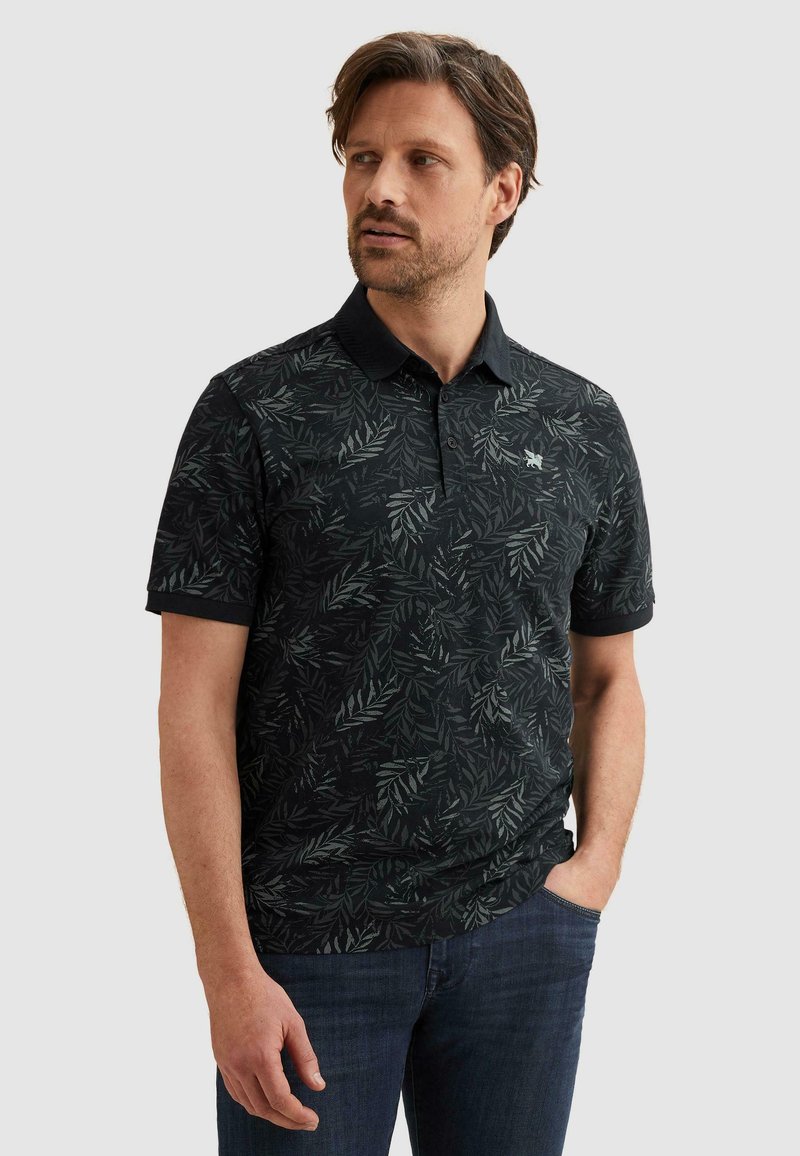 Homme portant un polo noir à manches courtes avec un motif feuillu foncé et un jean bleu, debout avec une main dans la poche, regardant vers la gauche.