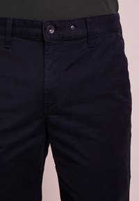 Pantaloni di cotone blu navy con chiusura a bottone, cuciture visibili e design frontale piatto. Texture liscia e vestibilità dritta standard.