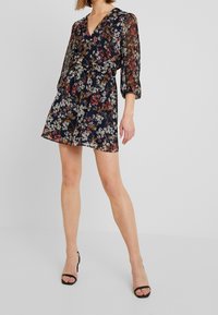 Mörkblå blommig omlotklänning med långa genomskinliga ärmar, med V-ringning och figursydd midja. Designen inkluderar färgglada blommönster och en utsvängd kjol.