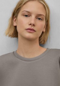 Camisetta grigia a maniche corte con scollatura rotonda realizzata in un tessuto liscio. Il modello ha i capelli lunghi fino alle spalle. Design semplice e minimalista.