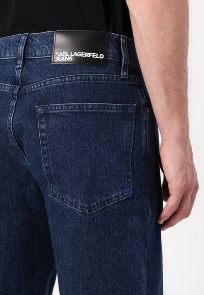 Jean en denim foncé à coupe droite, avec un patch en cuir étiqueté "Karl Lagerfeld Jeans" et un petit logo bleu sur la poche arrière.