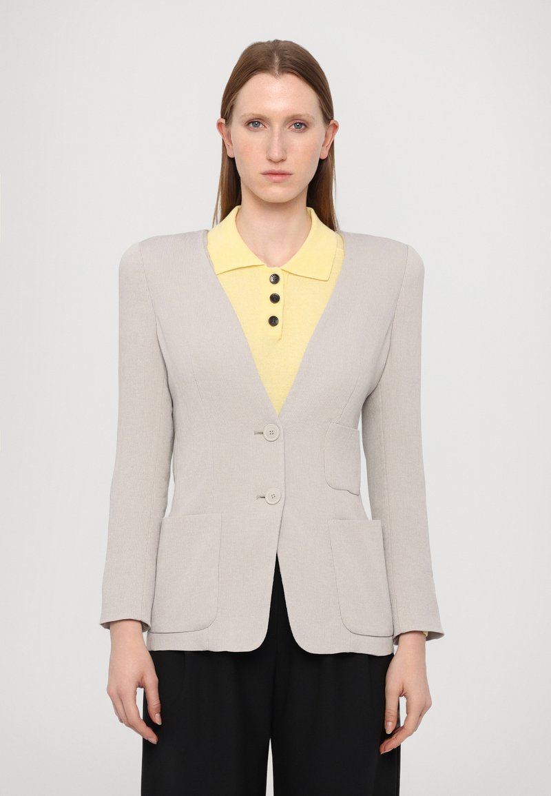Light grey blazer with a fitted design, two buttons, and two front pockets. Worn over a yellow collared shirt with black buttons.