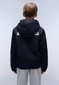 Schwarze Kapuzenjacke aus glattem Stoff mit elastischen Bündchen und einem Logodetail auf dem Rücken. Kombiniert mit hellgrauen Hosen.