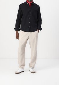 Sort langærmet skjorte med knaplukning og enkelt lomme, kombineret med lyse beige teksturerede bukser og hvide sneakers med grå detaljer.