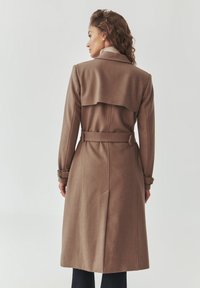 Brauner Wolltrenchcoat mit doppelreihigem Design, hohem Kragen, tailliertem Gürtel und Rückenkappe. Verfügt über lange Ärmel mit verstellbaren Manschetten.
