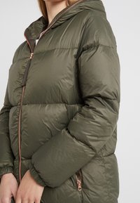 Nafouknutá olivově zelená bunda s kapucí, která má dvoucestný zip, elastické manžety a boční kapsy s akcenty v barvě růžového zlata. Hladká textura.