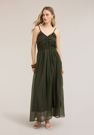 Junge Frau trägt ein olivgrünes, gerafftes Maxikleid mit Spaghettiträgern, beigen Sandalen, goldenen Ohrringen und braunen Armreifen, steht vor einem schlichten Hintergrund.