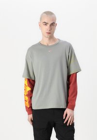 T-shirt oversize a maniche corte grigio, indossato sopra una maglietta rossa a maniche lunghe con grafica gialla. Tessuto morbido, collo rotondo, vestibilità casual.