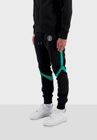 Černé tepláky s tyrkysovými bočními panely a elastickým pasem. Vyrobené z hladké látky; na přední straně mají logo. Nošené s bílými teniskami.