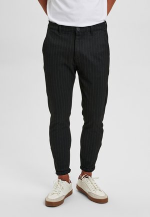 Man draagt zwarte smalle cropped broek met krijtstrepen en witte vetersneakers tegen een effen achtergrond.