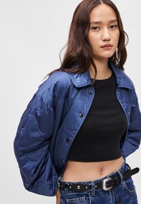 Μπλε επενδυμένο μπουφάν με γιακά, σκούρα κουμπιά και χαλαρή εφαρμογή, φορεμένο πάνω από ένα μαύρο τοπ και συνδυασμένο με τζιν ψηλής μέσης.