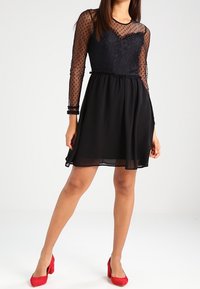 Black dress featuring a lace bodice with a sheer polka dot overlay, long sleeves, and a flowy skirt. Paired with red high heels.
