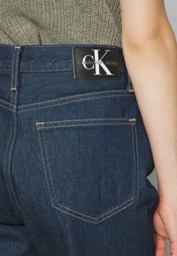 Vaqueros de mezclilla azul oscuro con una etiqueta negra que muestra el logo "CK", detalles de costura visibles y un diseño estándar de bolsillo trasero.