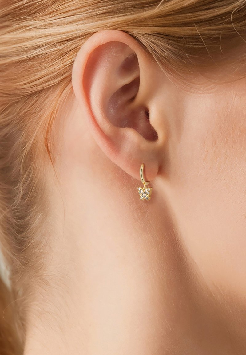 Zelta gredzenveida auskars ar tauriņa formas piekariņu, dekorēts ar maziem caurspīdīgiem akmeņiem, parādīts uz gaišas ādas un blondu matu fona.