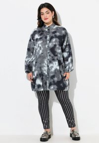 Overmaatse blouse in grijs en zwart tie-dye patroon, met lange mouwen en een kraag. Gecombineerd met zwart-grijze gestreepte leggings.