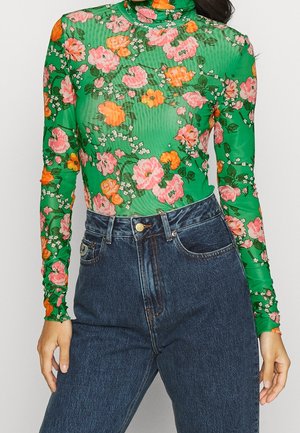 Grüner Langarm-Rollkragenpullover mit pinkem und orangefarbenem Blumenmuster, kombiniert mit hoch taillierten dunkelblauen Jeans an einer stehenden Person.