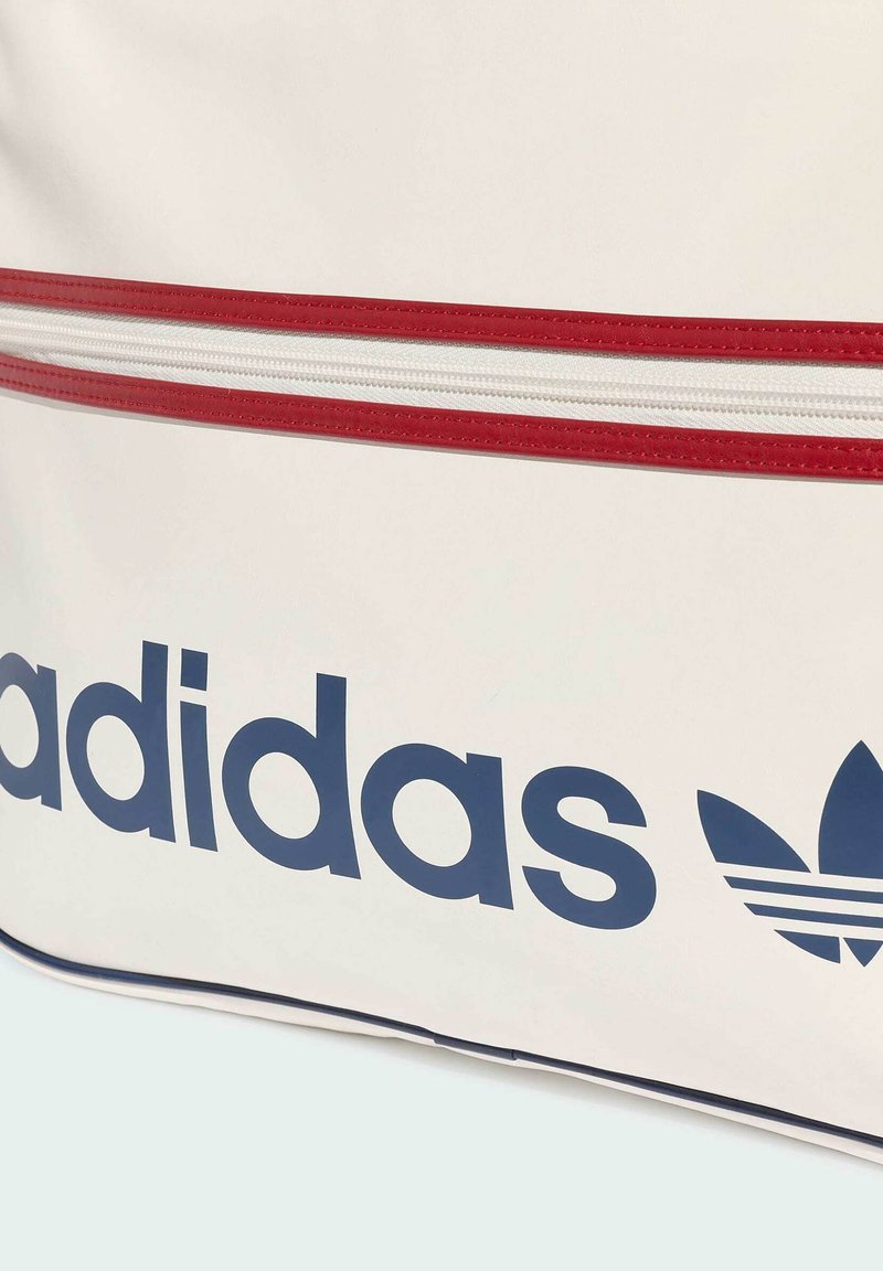 Sezione di borsa bianca con striscia della cerniera rossa e logo Adidas blu navy con simbolo trifoglio su sfondo a superficie liscia.