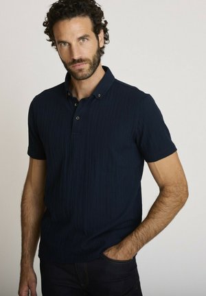 Homme aux cheveux bouclés et à la barbe, portant un polo bleu marine à manches courtes, debout avec une main dans la poche sur un fond uni.