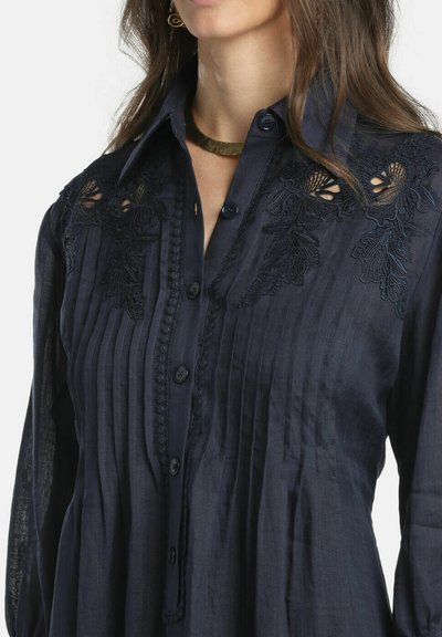 Žena v tmavomodrej blúzke na gombíky s vertikálnymi záhybmi a čiernou čipkovou výšivkou na pleciach, doplnená zlatým obojkom.