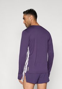 Lila Langarmshirt mit weißen Seitenstreifen und goldenem "ADIZERO"-Logo; getragen mit passenden Shorts aus strukturiertem Stoff und seitlichen Schlitzen.