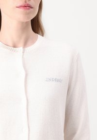 Krémový kardigan vyrobený z měkké pleteniny, s žebrovaným límcem, zapínáním na knoflíky a malým logem "2ND DAY" ve světle modré barvě.