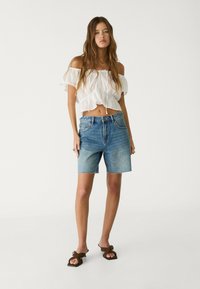 Kevyt valkoinen off-shoulder-toppi, jossa on röyhelöitä, yhdistettynä sinisiin denimshortseihin, joissa on revitty helmastus ja rento istuvuus. Ruskeat sandaalit täydentävät asua.