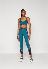 Donna in reggiseno sportivo Puma teal e nero e leggins, con scarpe bianche, davanti a uno sfondo bianco.