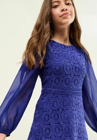 Abito blu in pizzo caratterizzato da un motivo testurizzato, lunghe maniche trasparenti e scollatura rotonda con un corpetto aderente e dettagli arricciati in vita.