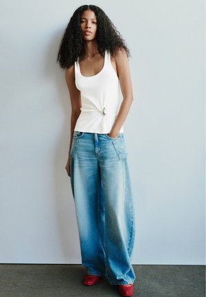 Femme aux longs cheveux bouclés portant un haut blanc sans manches, un jean large bleu clair et des chaussures rouges, debout contre un mur uni.