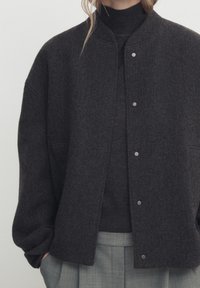 Veste en mélange de laine gris foncé avec un col rond, des fermetures à pression et une coupe décontractée. Portée sur un pull assorti de couleur sombre, associée à un pantalon gris clair.